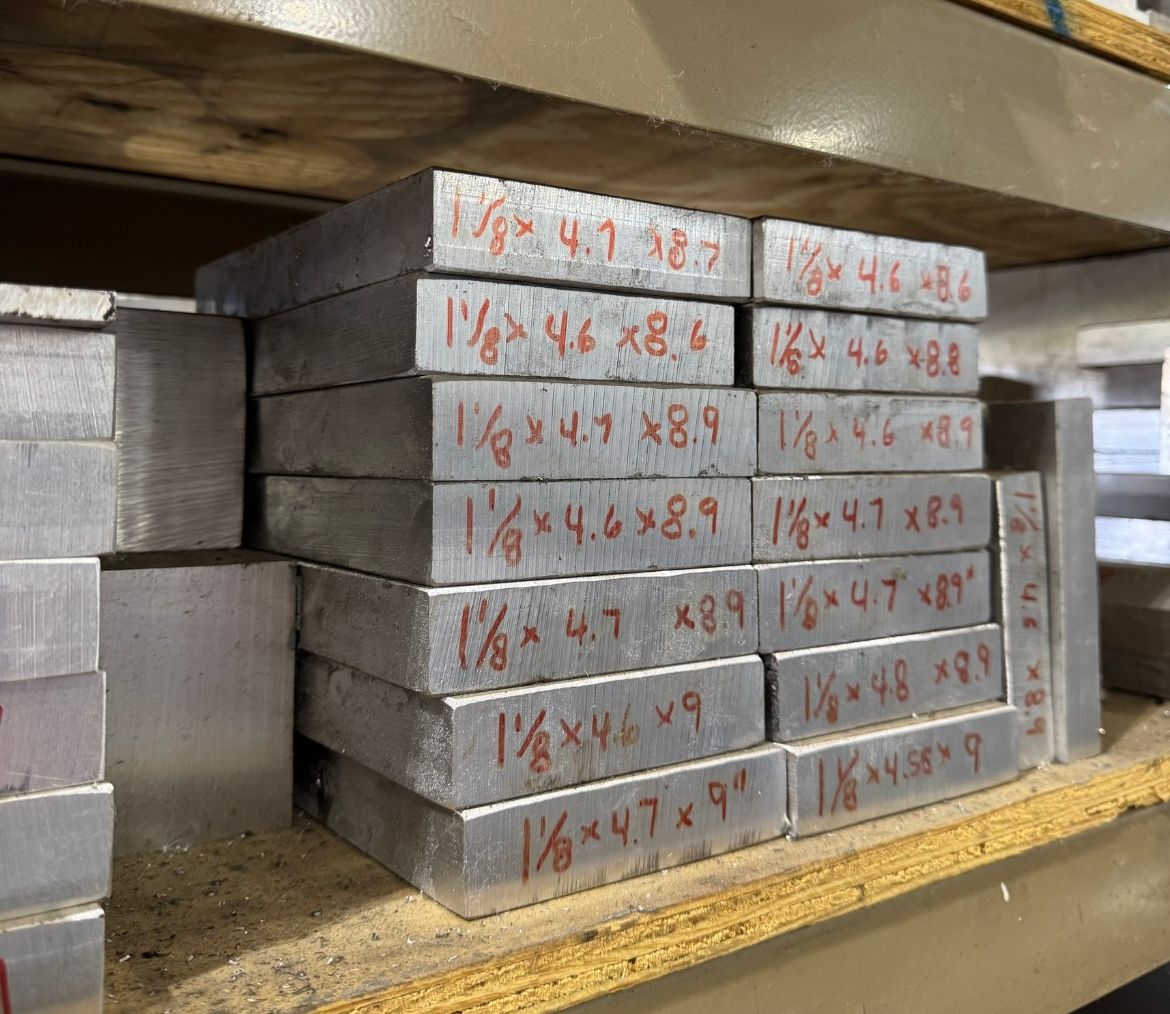 Stack of metal rectangular bars with handwritten size markings in red on their ends, organized on a shelf.