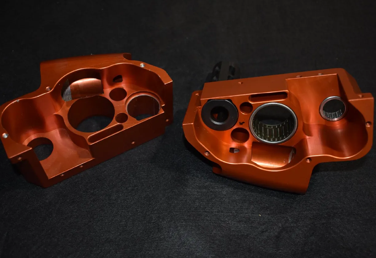 Two orange anodized metal machining components with circular cutouts and bearing inserts on a black surface.