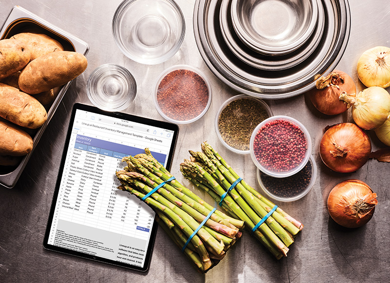 Ingredients for cooking on stainless steel countertop next to an ipad with inventory information listed.