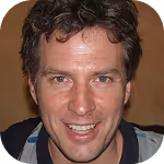 Smiling man with curly dark hair and blue eyes wearing a black and blue jacket.