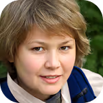 Smiling young person with short light brown hair wearing a blue and beige jacket outdoors.