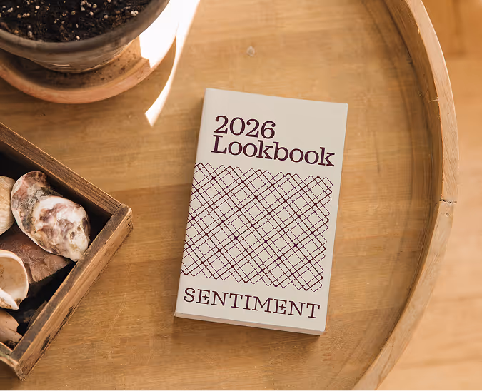 2026 Lookbook Sentiment book on a round wooden table beside a wooden box with shells and a plant pot.