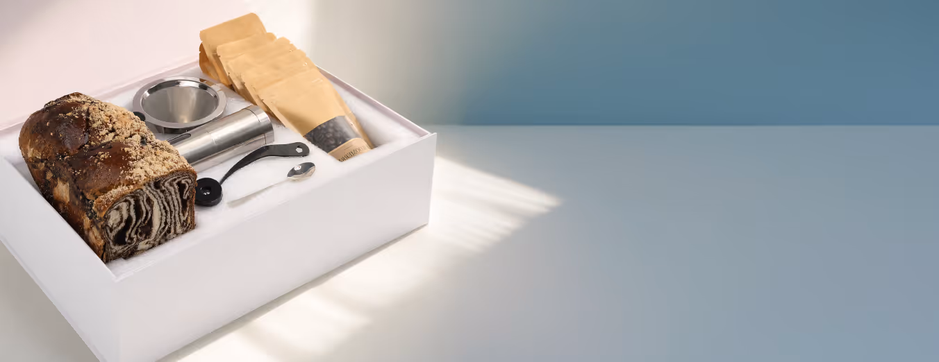 Gift box containing a loaf of marble bread, metal funnel, coffee measuring spoon, and brown coffee bean bags.
