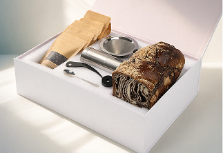 White gift box containing a loaf of patterned bread, metal scoop, black spoon, and three brown paper bags.