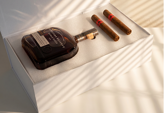 Bottle of dark liquor and two cigars resting in a cushioned display box with striped light shadows.