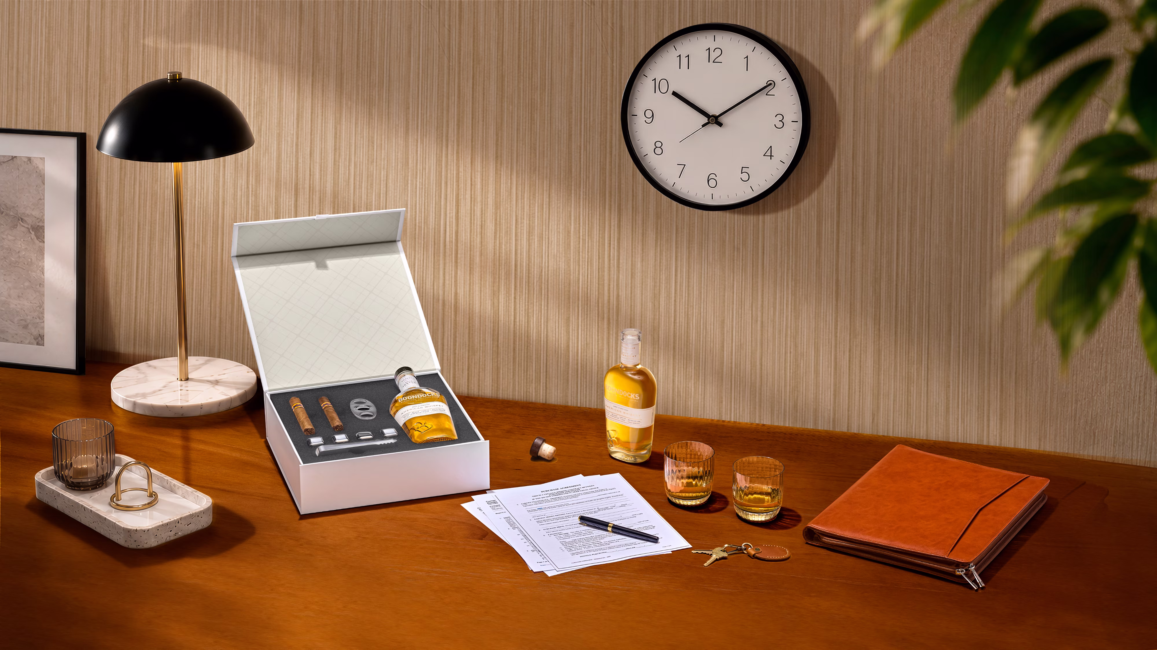 Wooden desk with a lamp, open box with whiskey and cigars, two glasses, documents, keys, and a leather folder.