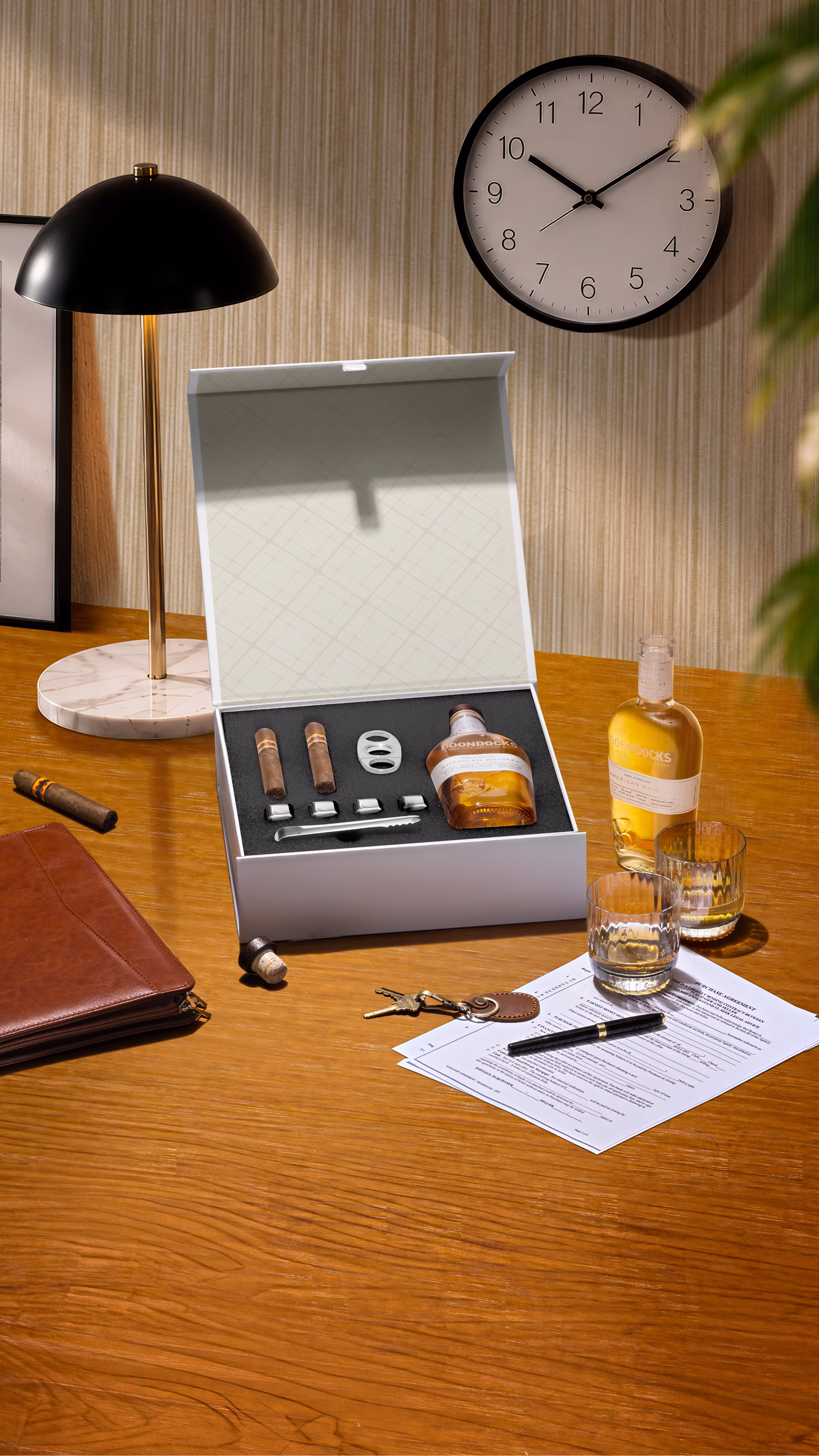 Wooden desk with cigars, whiskey bottle and set, glasses, papers, pen, keys, and modern lamp under clock.