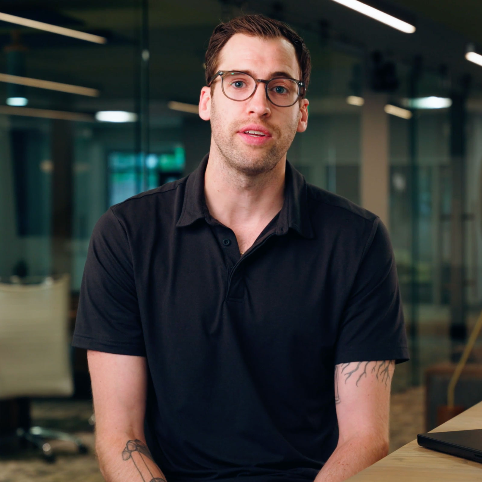 Man in glasses and black polo shirt sitting in an office with a laptop on a desk.