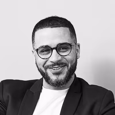 Bearded man with glasses smiling, wearing a black blazer and white shirt against a light background.