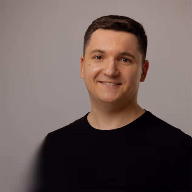 Smiling man with short dark hair wearing a black shirt against a neutral background.