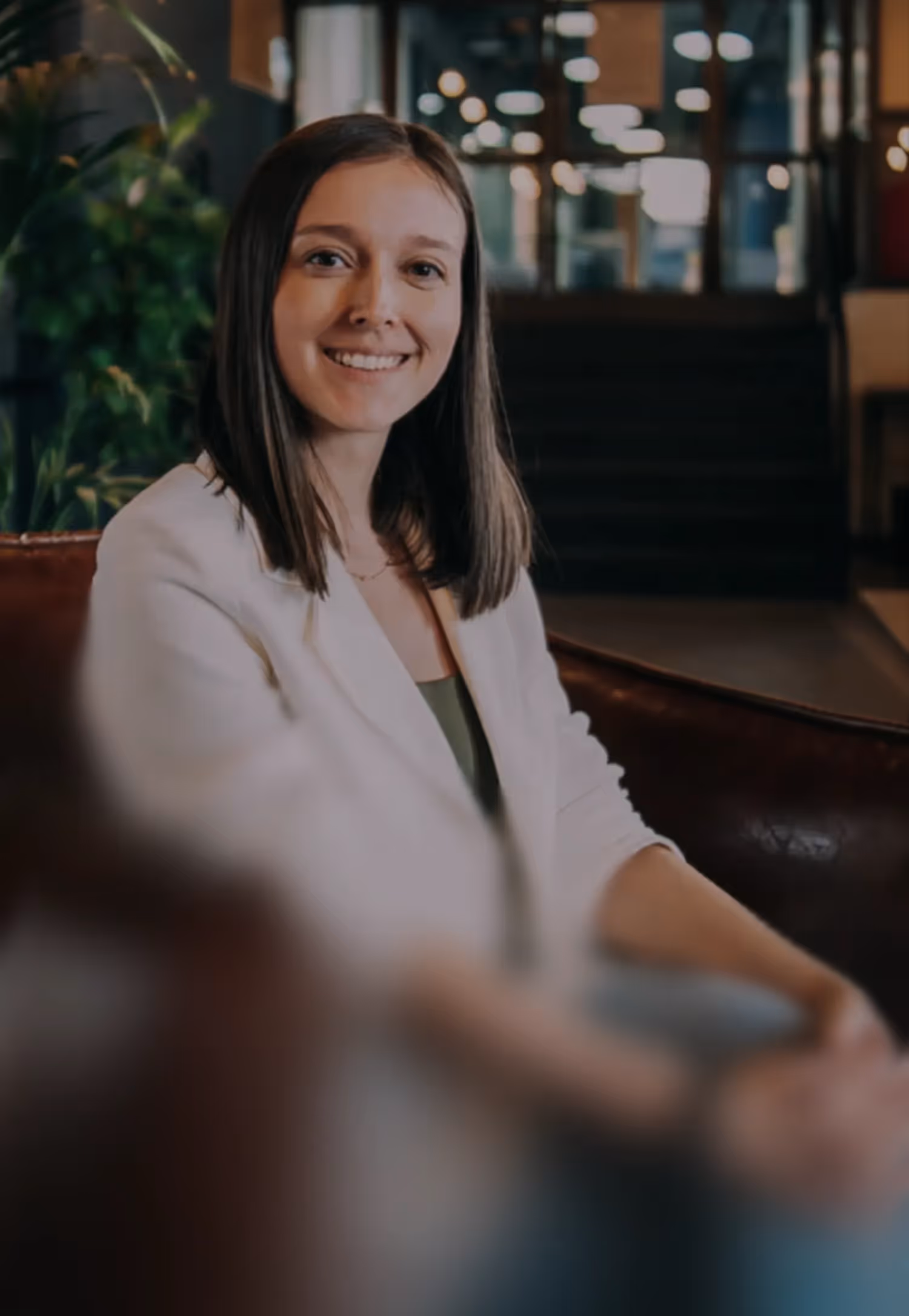 Smiling woman with shoulder-length brown hair wearing a white blazer sitting on a brown leather sofa indoors.