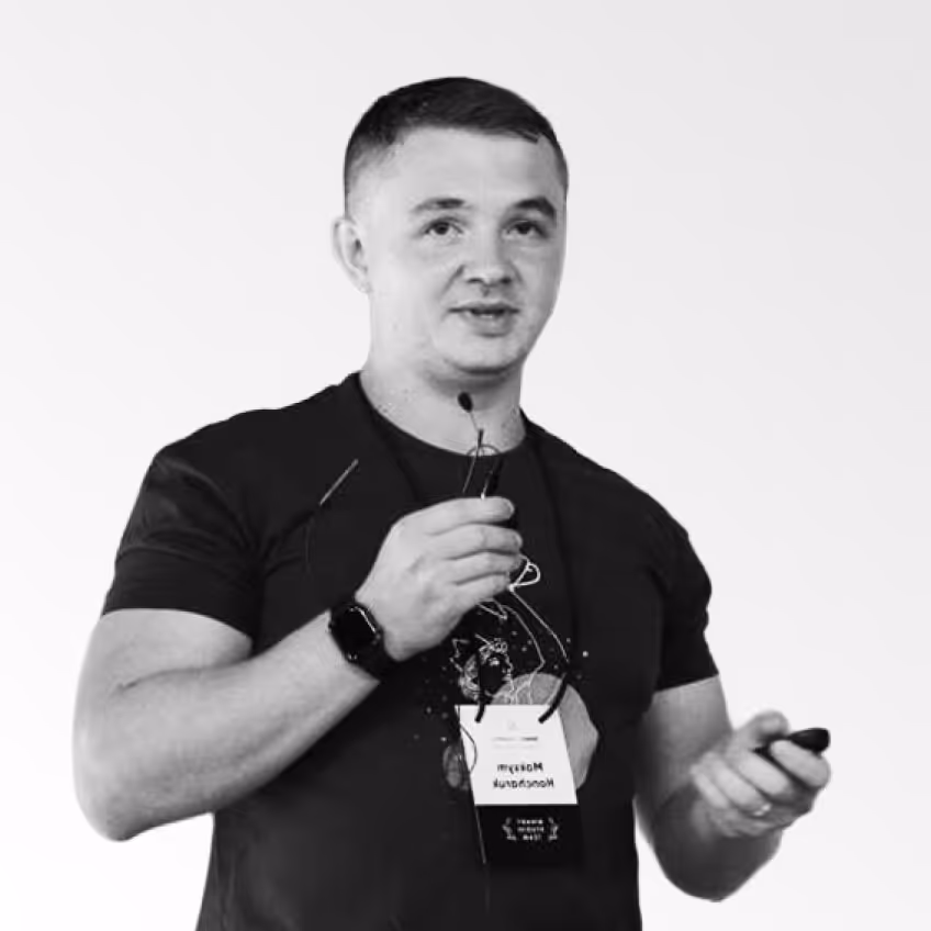 Man in a black t-shirt holding a microphone and a remote, speaking in front of a white background.