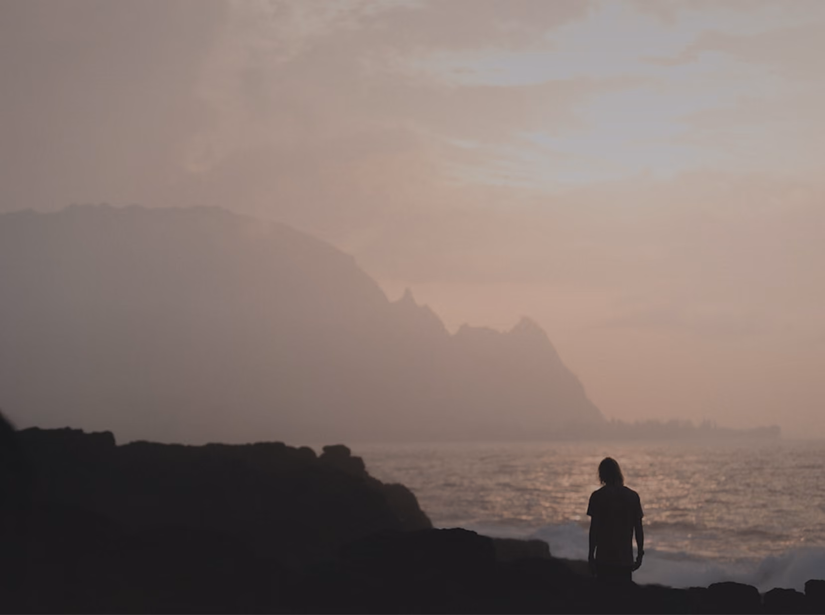 Silhouette of a person standing on rocky shore looking out at misty mountains and ocean at dusk.