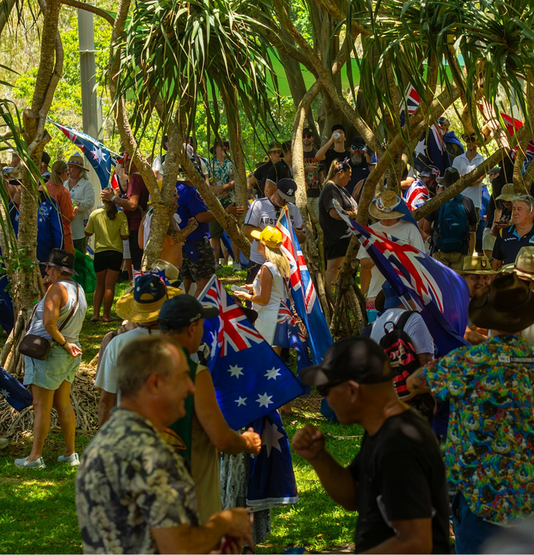 Crowd gathering outdoors under trees holding Australian flags in a sunny park setting.