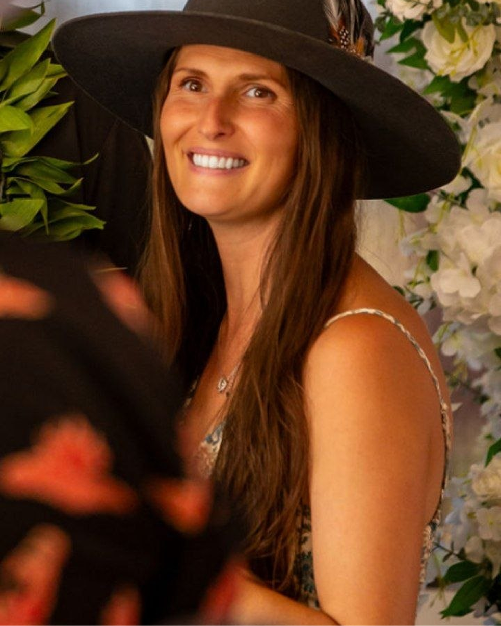 Smiling woman with long brown hair wearing a large dark hat and floral dress near white flowers.