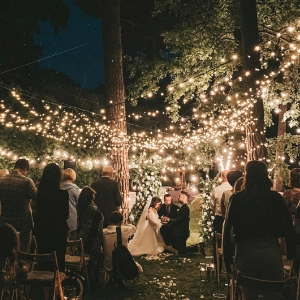 Outdoor evening wedding ceremony with bride and groom under string lights and surrounded by guests.