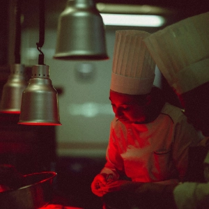 Two chefs in white uniforms and hats working together in dimly lit kitchen with red lighting.