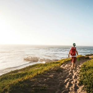 Person with a red backpack walking on a sandy path along a grassy coastline by the ocean at sunset.