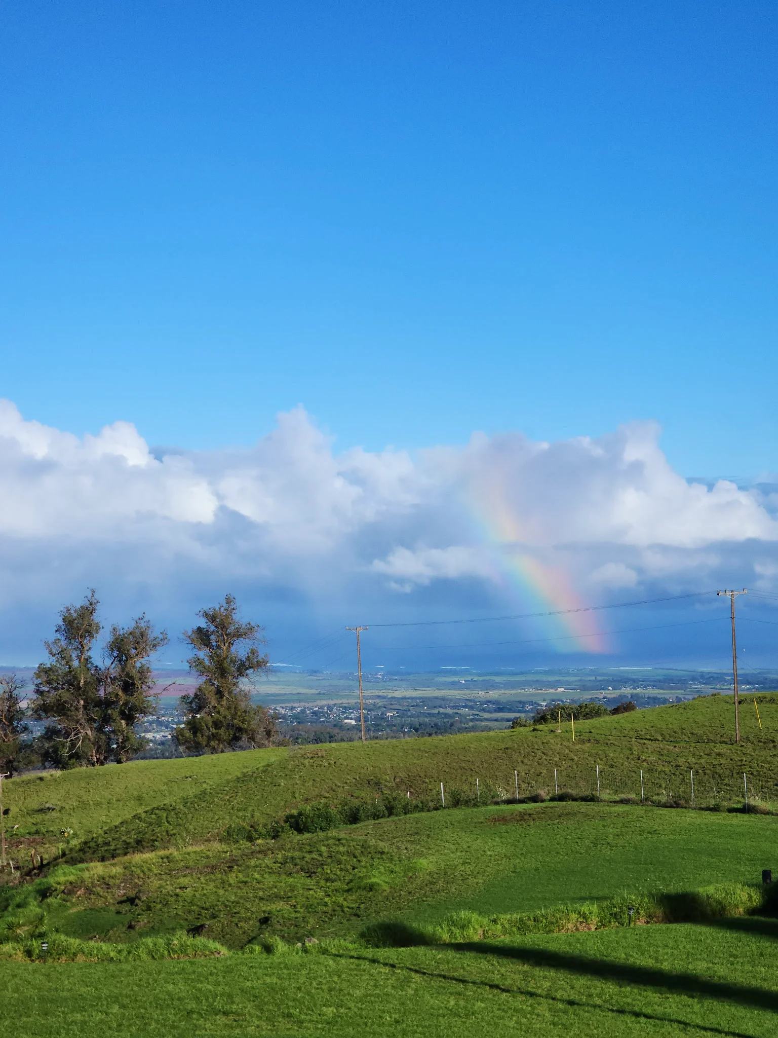 Green rolling hills under a blue sky with clouds and a faint rainbow over a distant town.