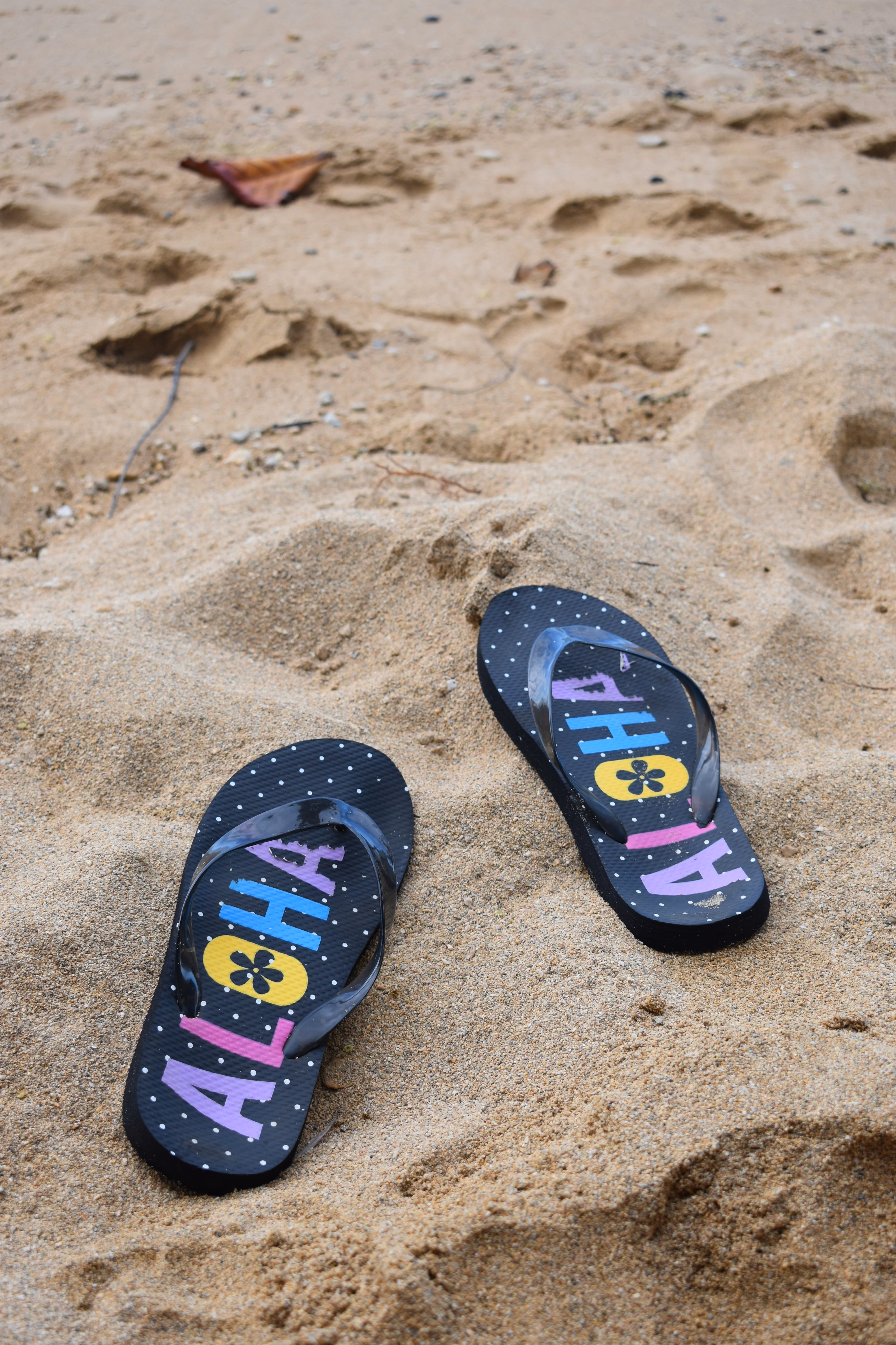 Black flip-flops with colorful ALOHA text and polka dots on sandy beach with footprints.
