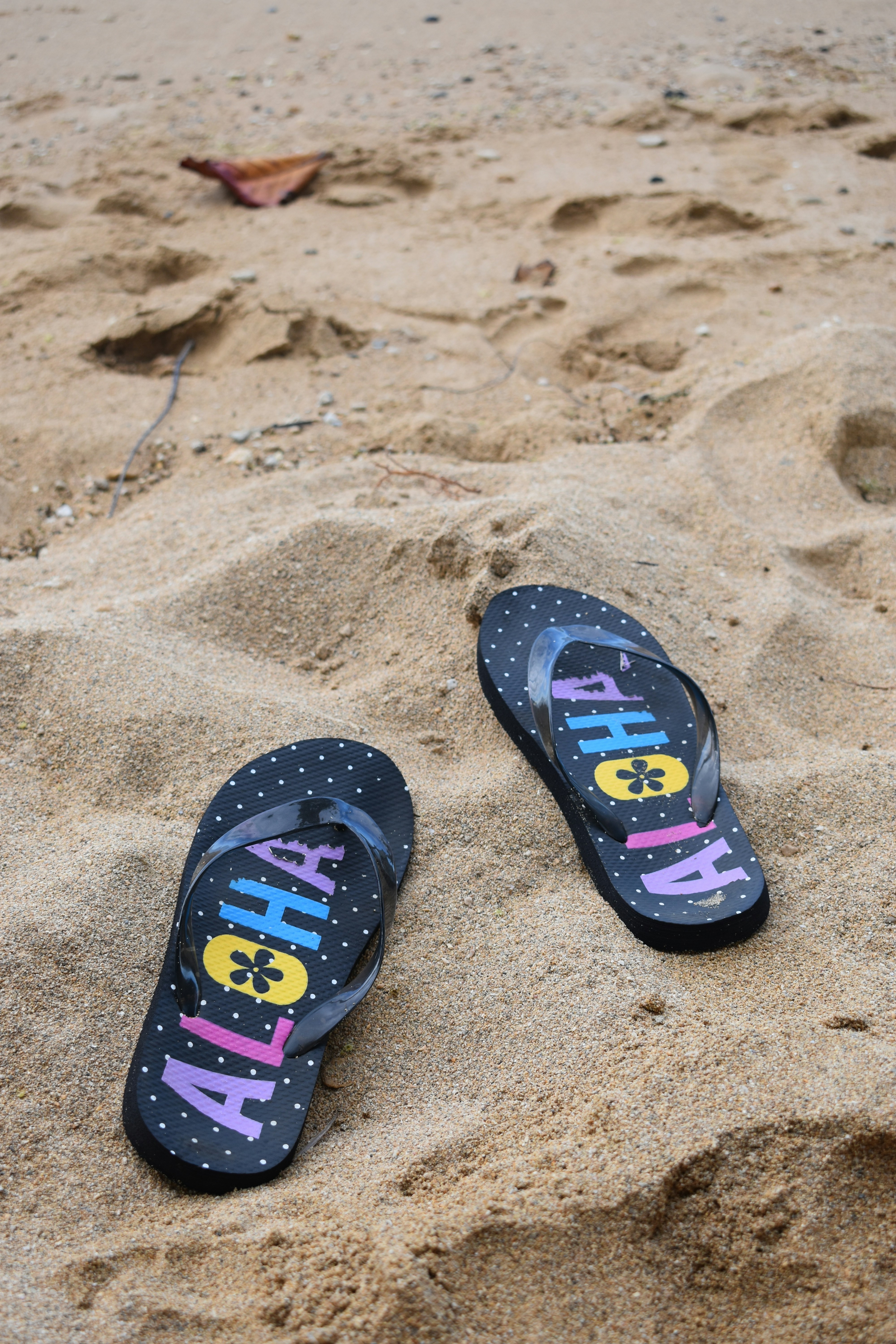 Black flip-flops with colorful ALOHA text and polka dots on sandy beach with footprints.