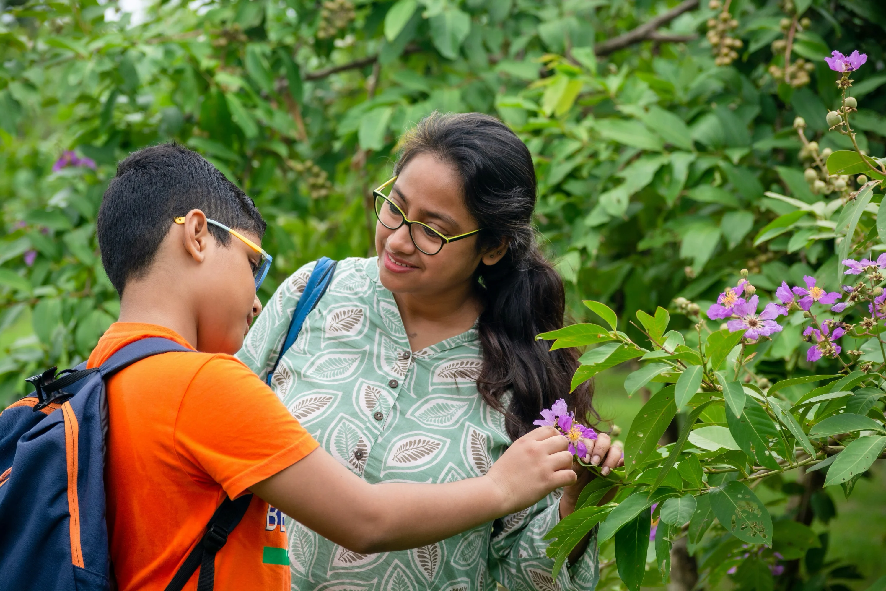 Mother and son examining a purple flower together in a green garden or park.