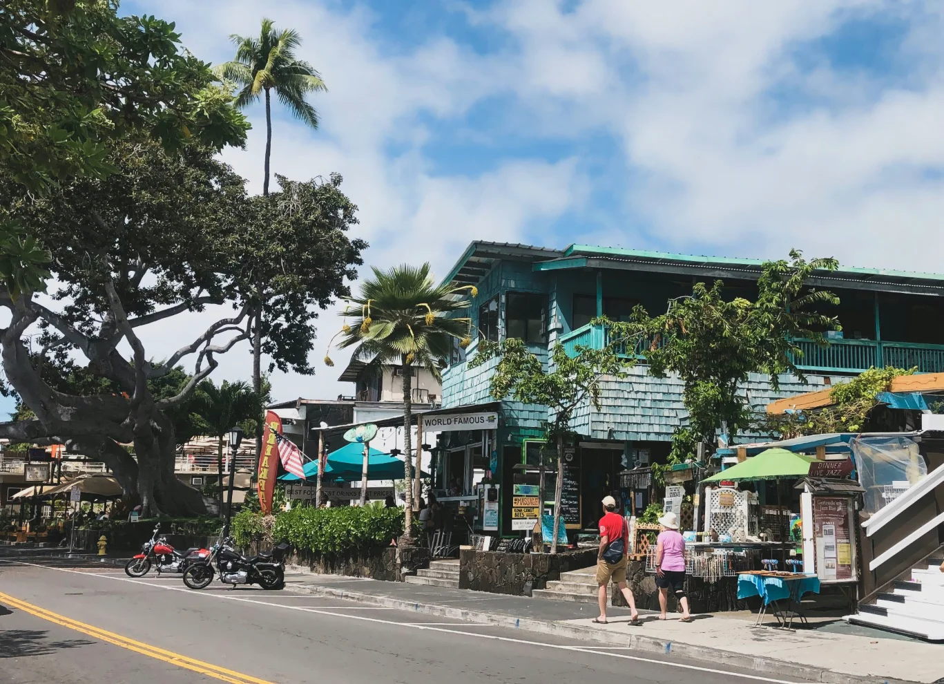 Street view of turquoise wooden building with shops, palm trees, and two pedestrians walking on the sidewalk.