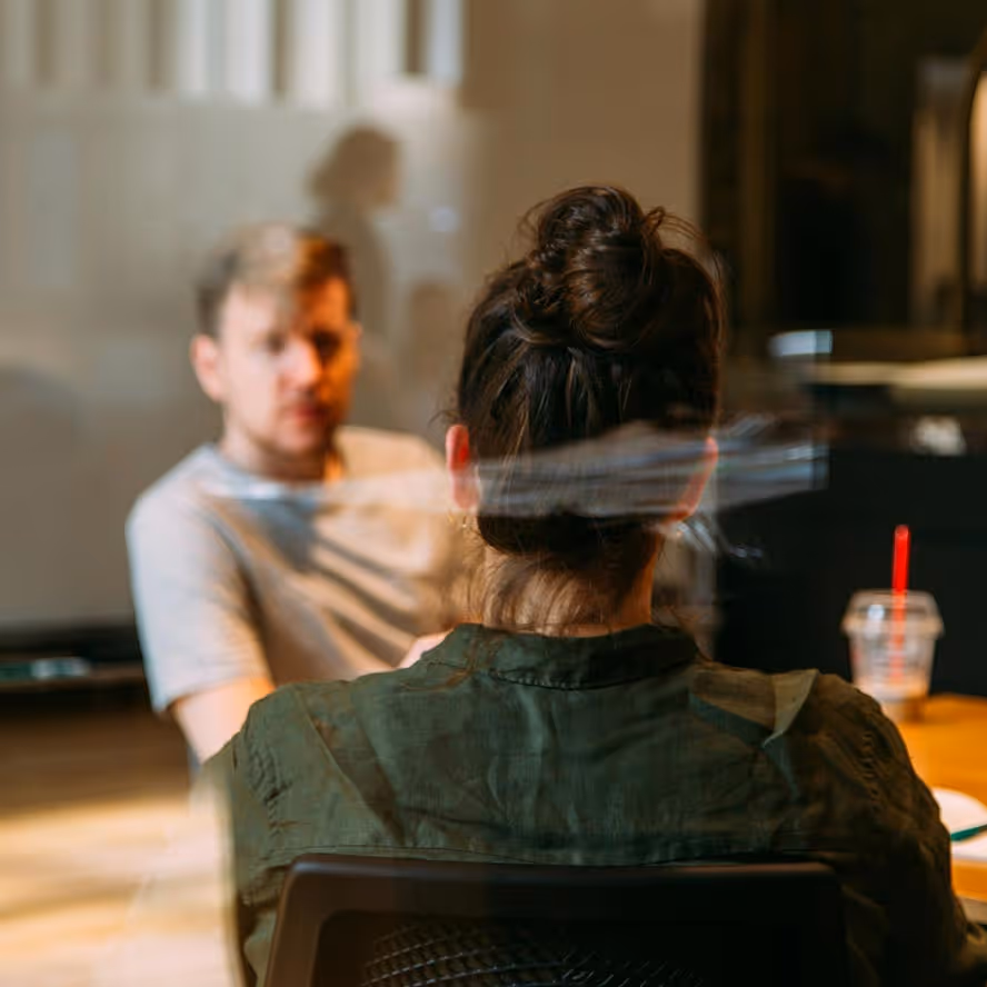 Two people having a conversation across a table in a softly lit room, with the woman’s back toward the camera and the man facing forward.