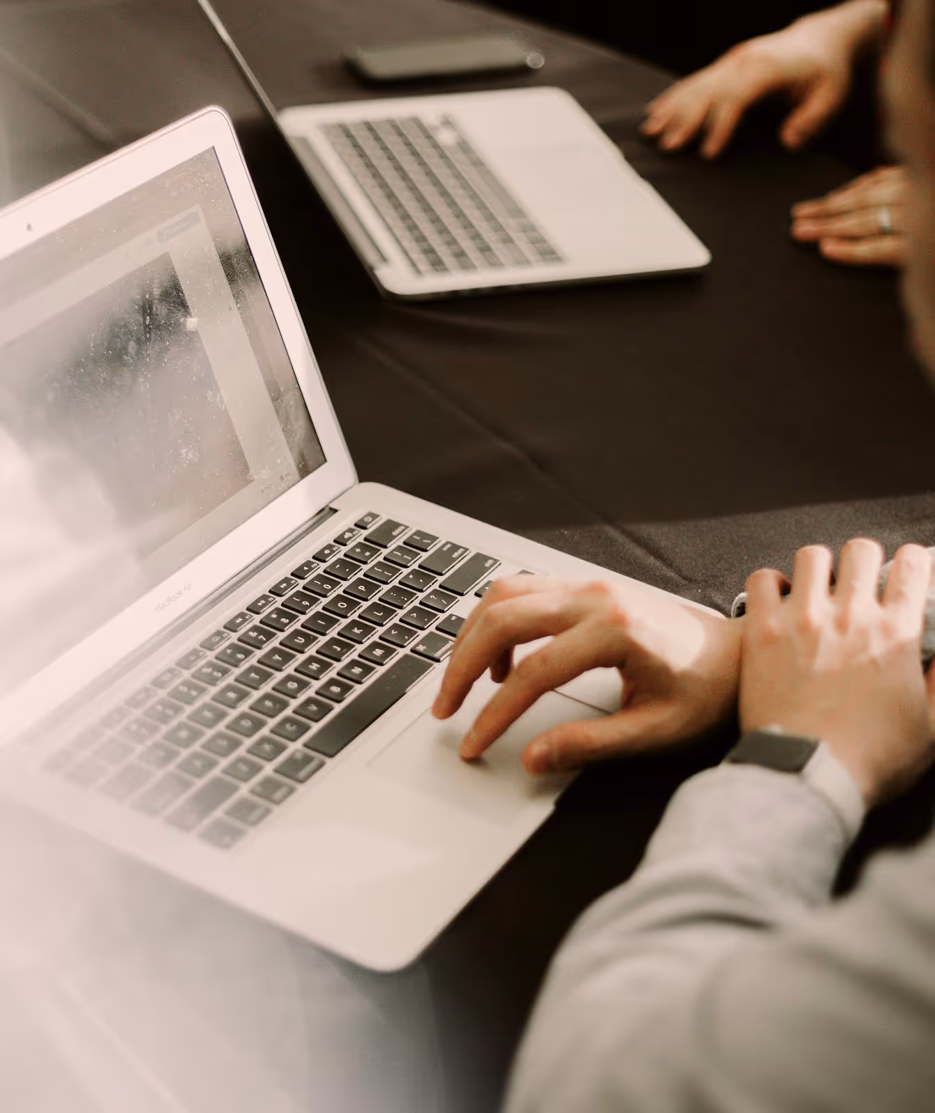 Person using a laptop on a dark table with another closed laptop and a smartphone nearby.