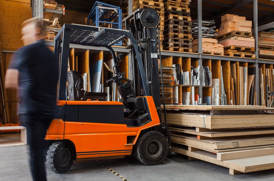 Orange forklift inside a warehouse with stacks of wood panels and a blurred person walking nearby.