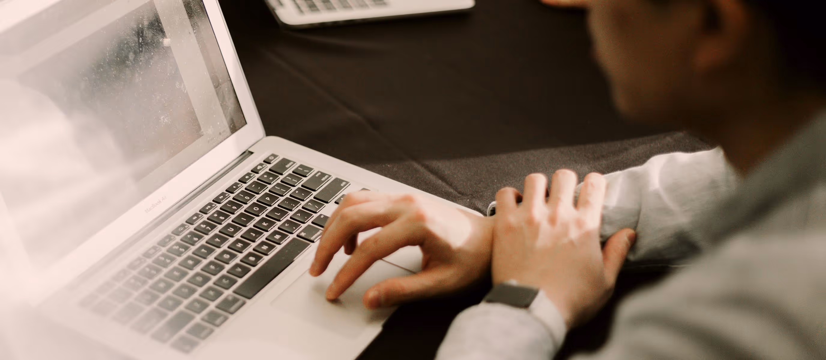 Person using a laptop on a dark table with another closed laptop and a smartphone nearby.