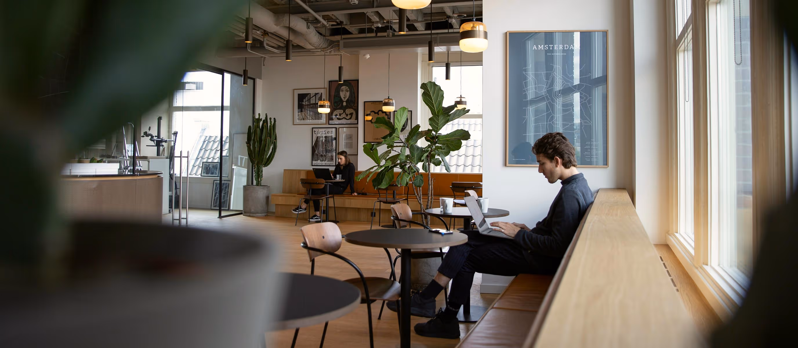 Two people working on laptops in a modern cafe with hanging lights, potted plants, and Amsterdam map artwork.
