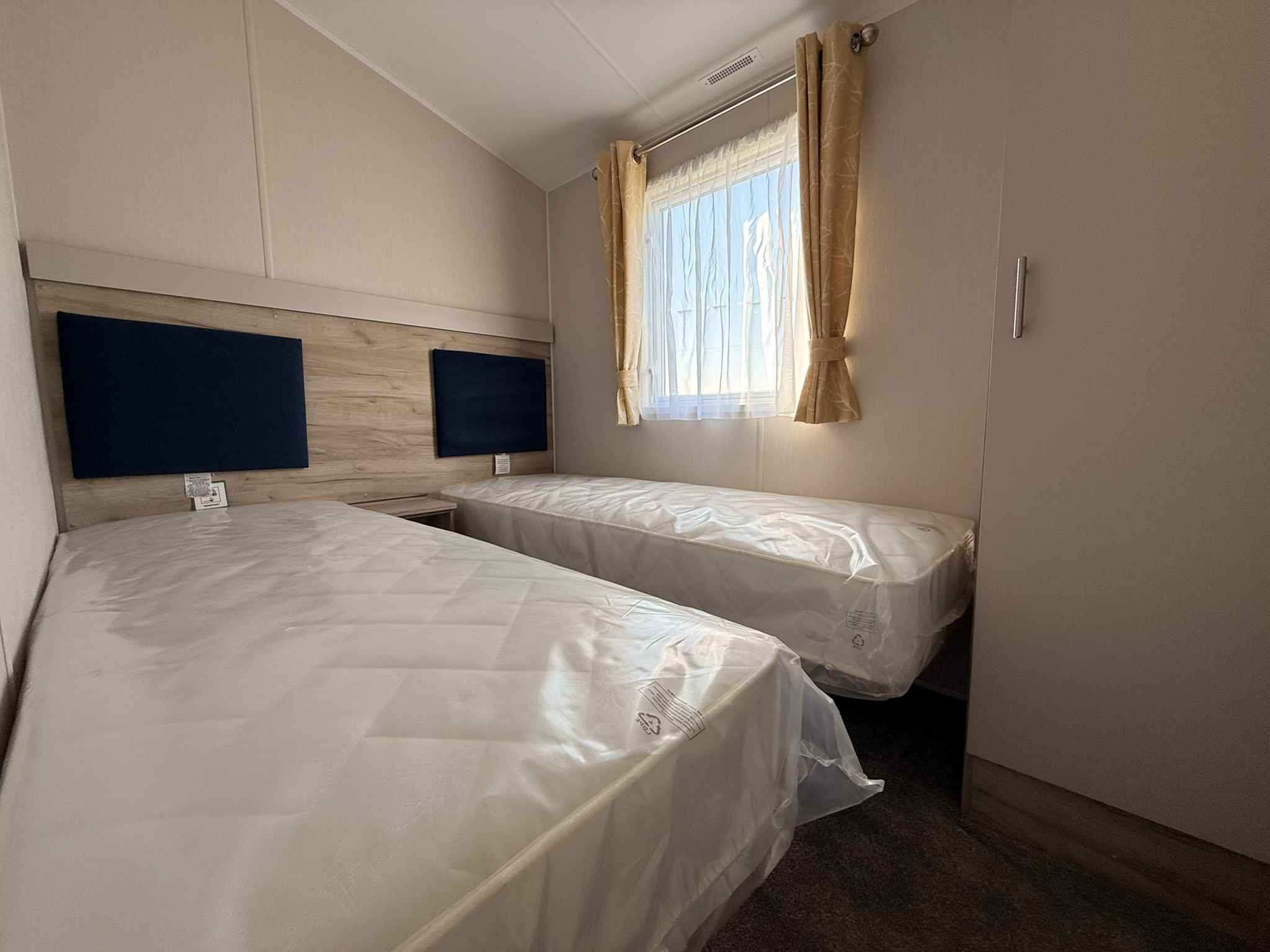 Modern hotel room with two single beds wrapped in plastic, mounted TV screens, and window with golden curtains.