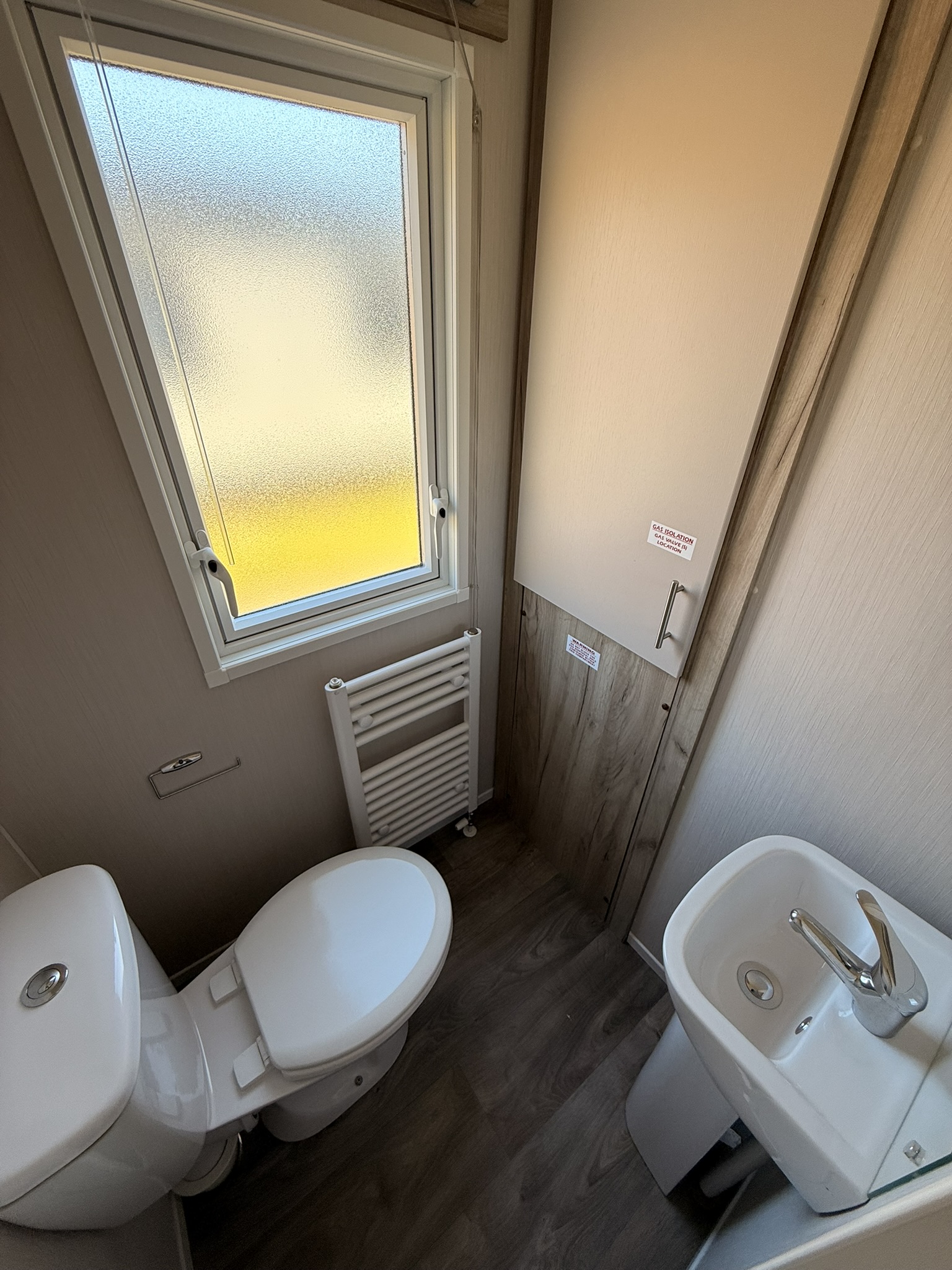 Modern bathroom with toilet, sink, radiator, and frosted window with yellow light outside.