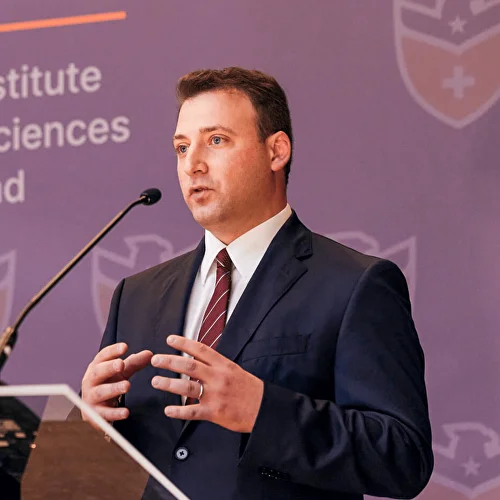 Man in dark suit and striped tie speaking at a podium with a microphone against a purple backdrop.