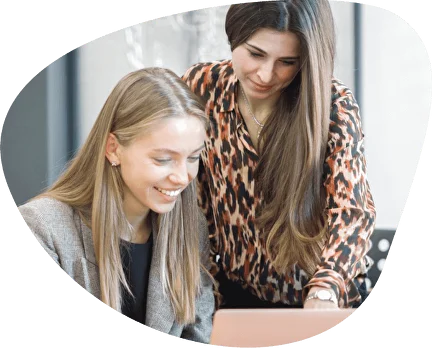 Two women smiling and looking at a laptop, seated in a bright indoor setting.