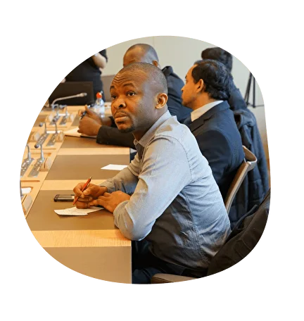 Man sitting at a conference table, writing on paper with a pencil, with others seated around him.