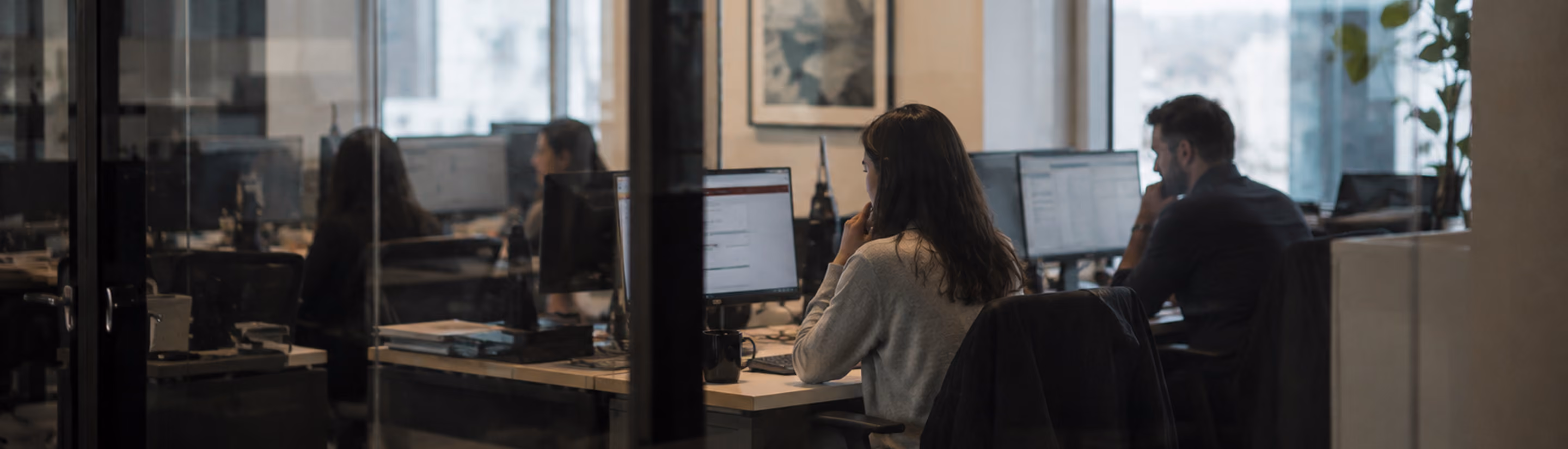 People working at desks on computers in a modern office with glass walls and large windows.