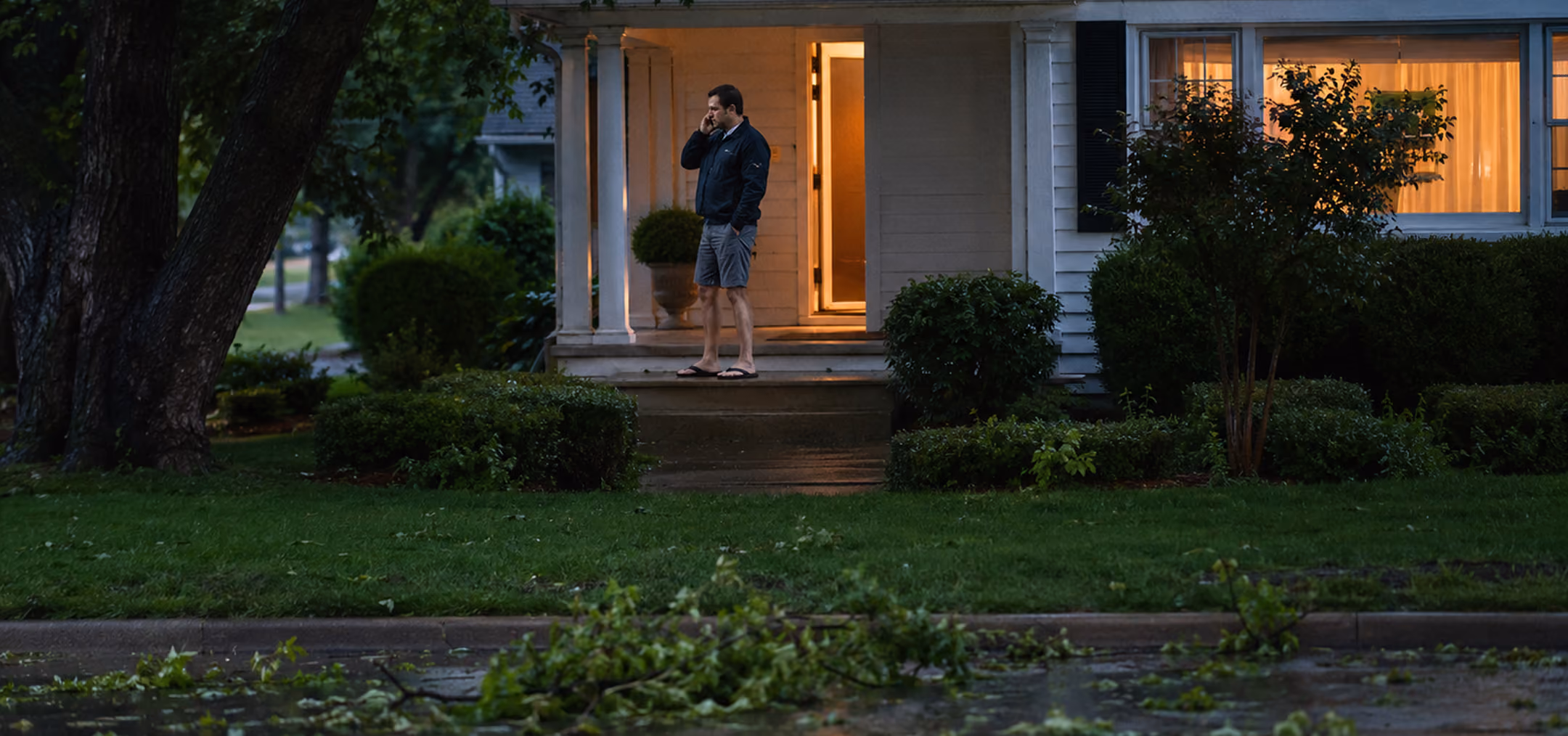 Man in shorts and jacket standing on a porch, talking on phone, with fallen branches on wet street.