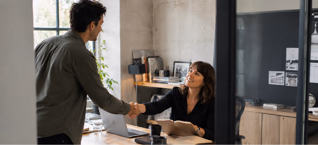 Woman sitting at desk shaking hands with man standing in modern office setting.