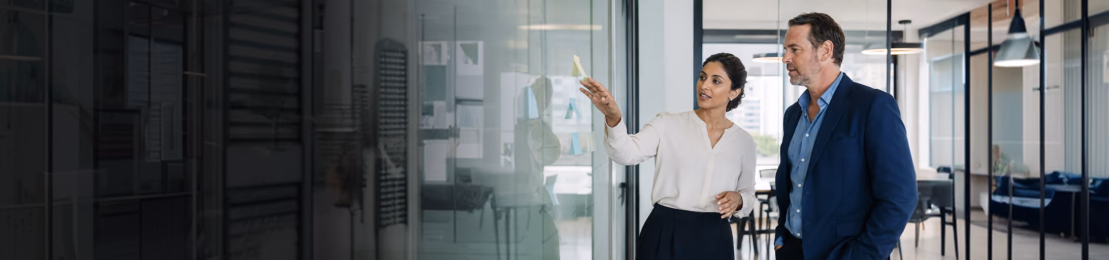 Two professionals discussing ideas while reviewing notes on a glass board in a modern office.