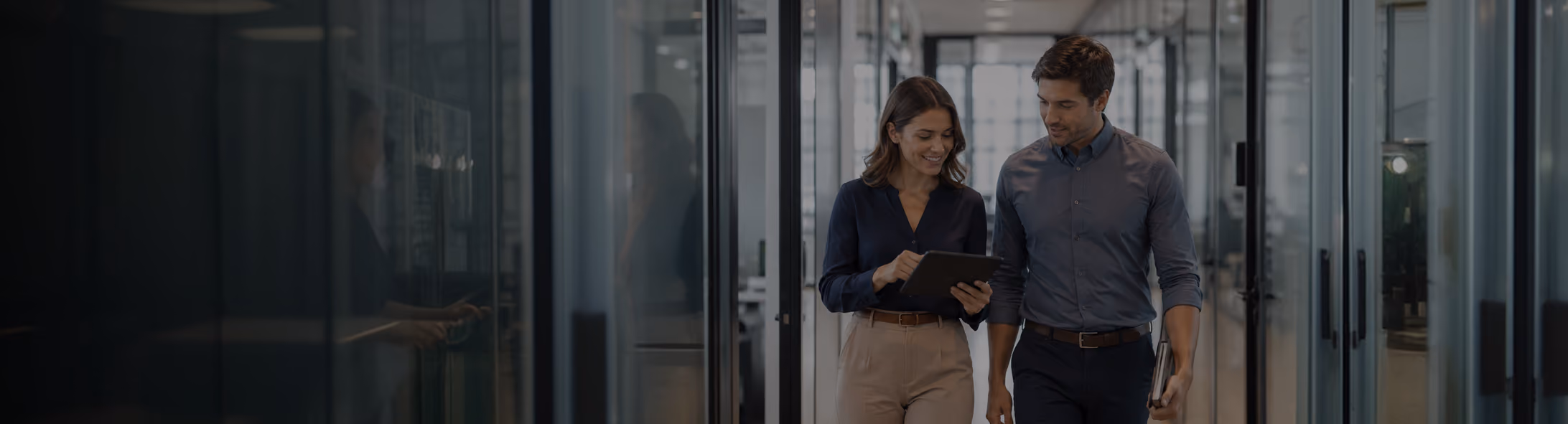Two colleagues walking through a modern office hallway while reviewing information together on a tablet.