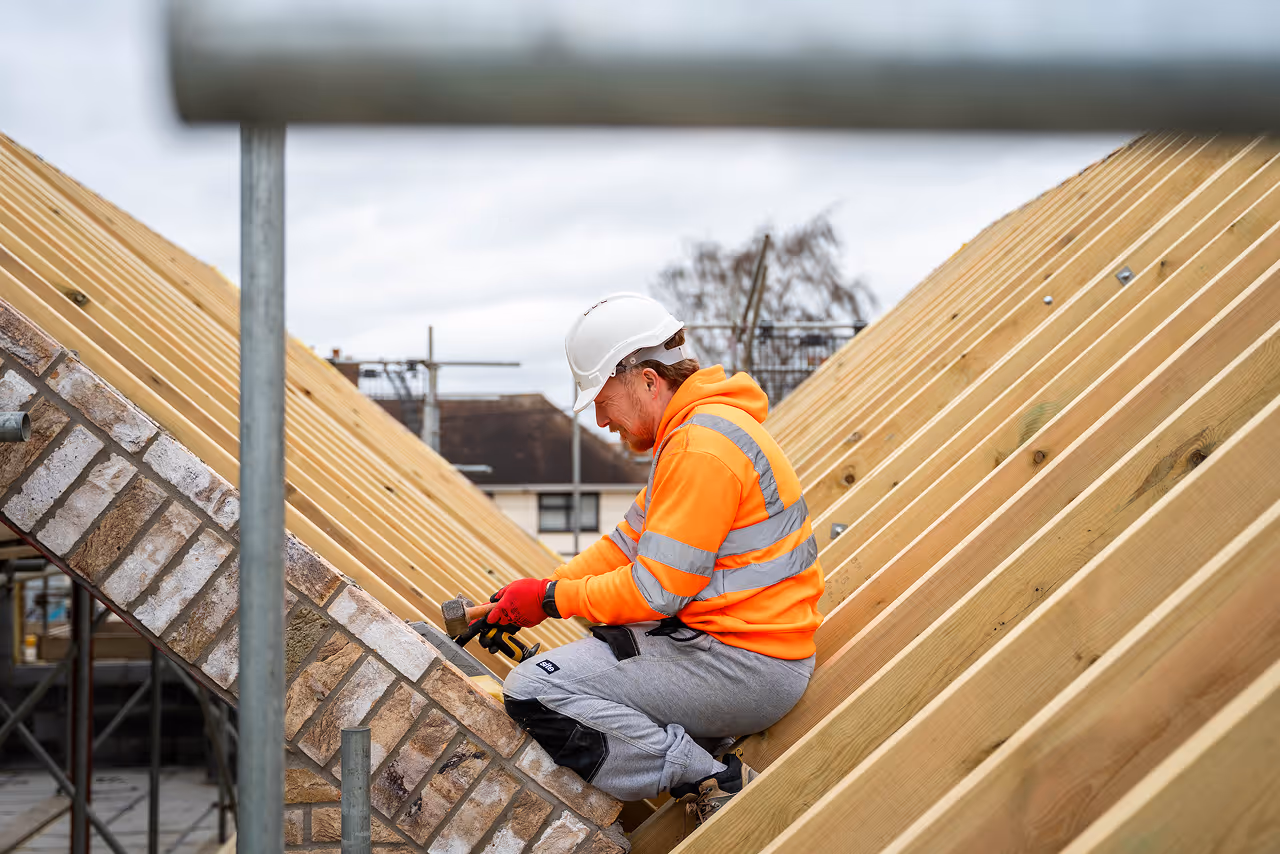A construction worker wearing a bright orange safety jacket and white hard hat works on a sloped roof. He uses tools with focus and precision.