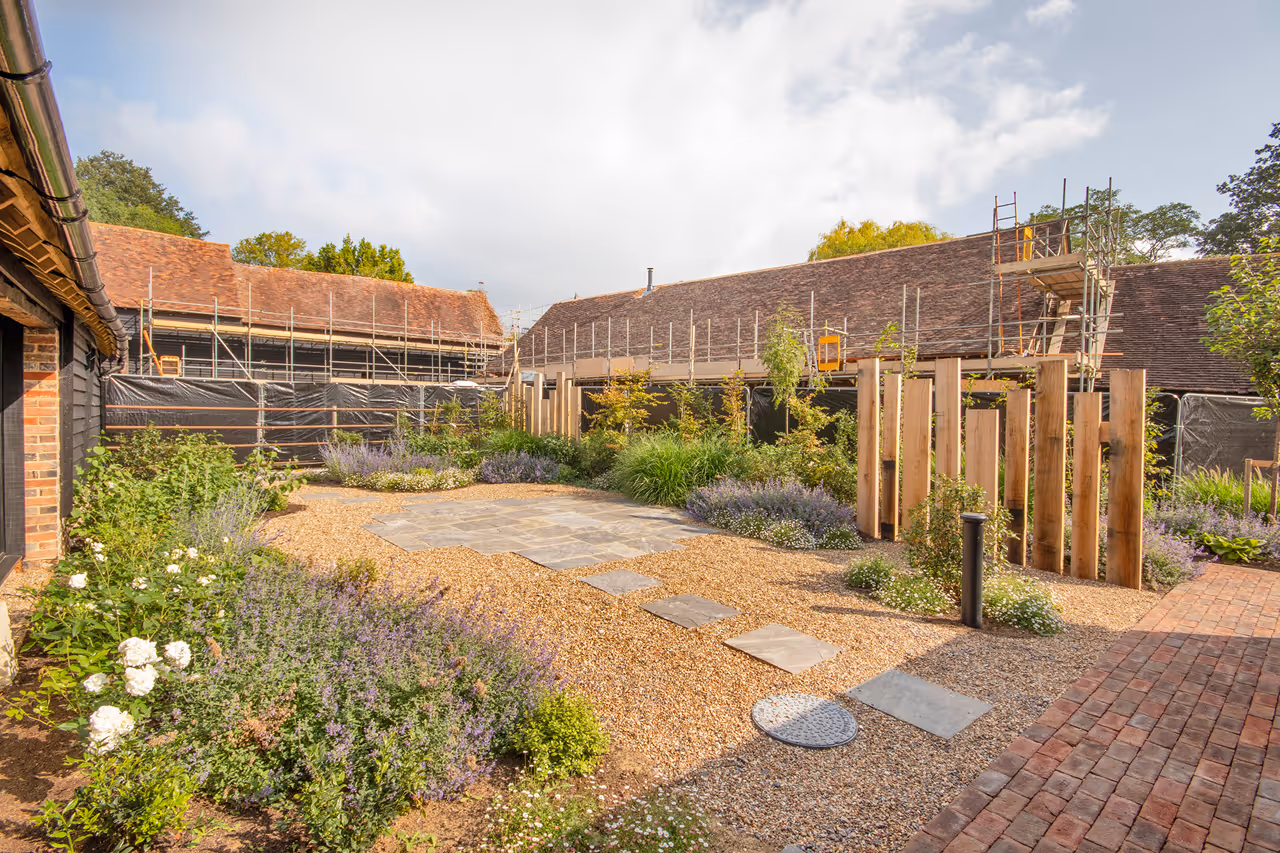 A tranquil courtyard with a mix of gravel and stone pathways, surrounded by lush greenery and flowers. Buildings under construction frame the scene, conveying a blend of nature and development.