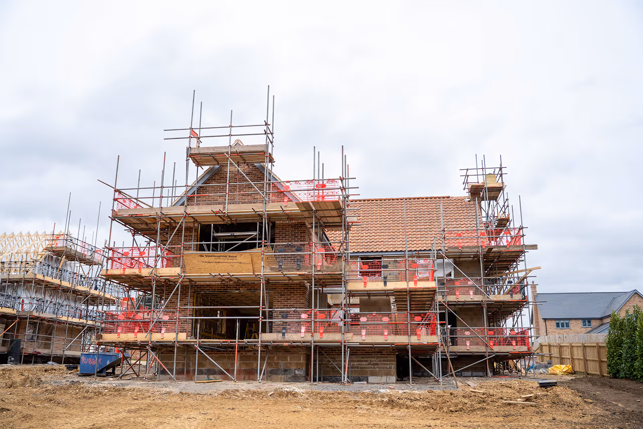 Partially built brick house with scaffolding around it on a construction site under cloudy sky.