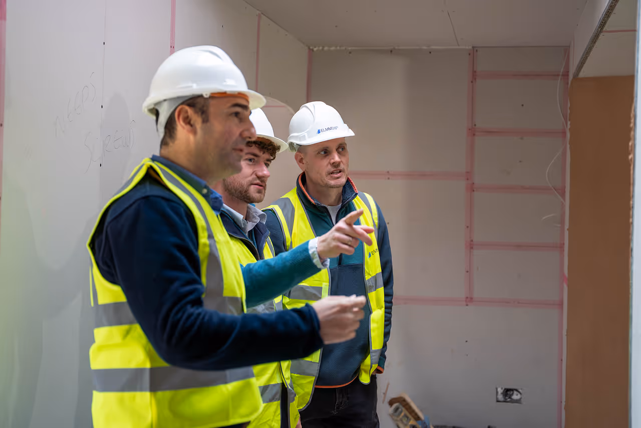 Three construction workers in hard hats and vests discussing plans inside a building under construction.