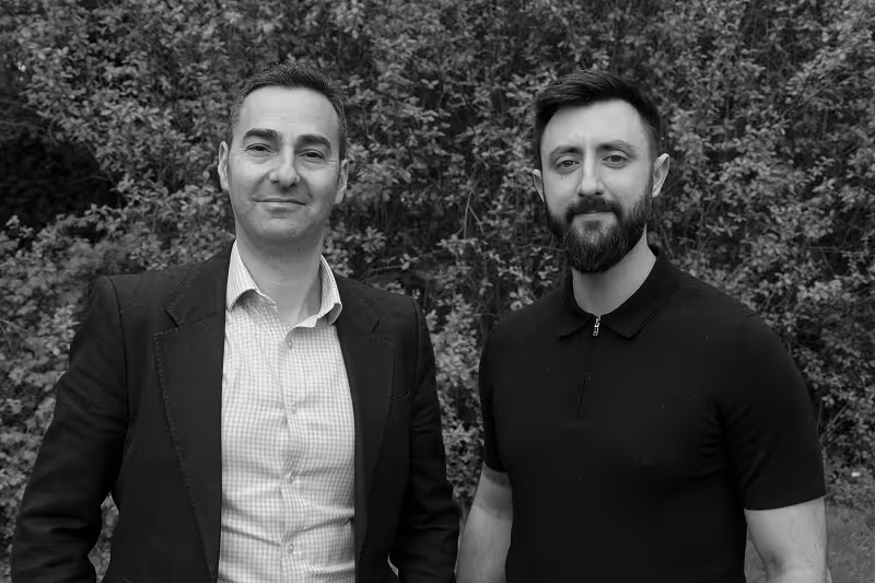 Two men standing outdoors in front of dense foliage, one in a suit jacket and one in a black polo shirt.