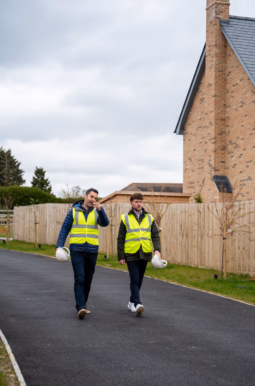 Two men in reflective vests holding white hard hats walking on a paved road beside wooden fences and brick houses.
