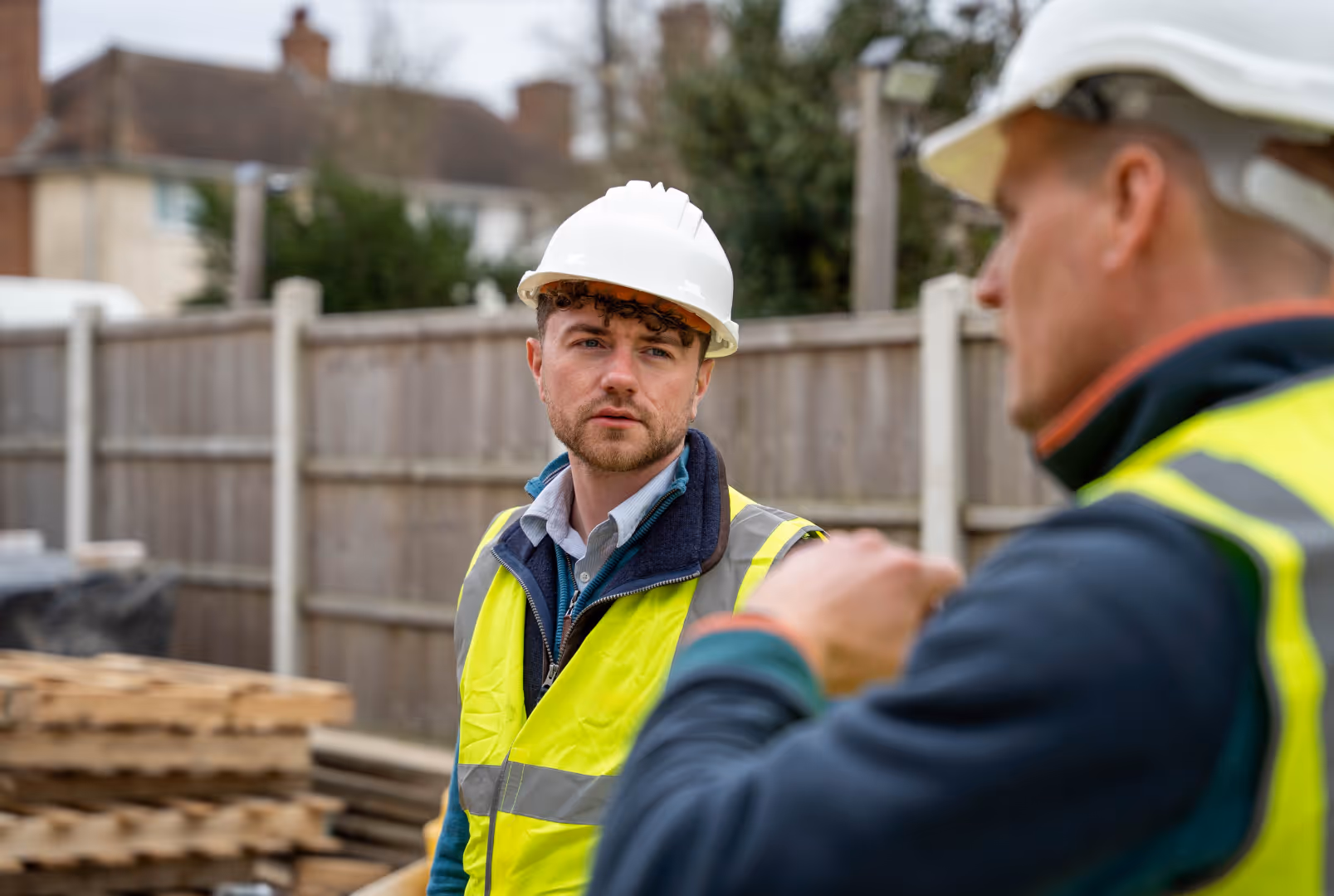 Two construction workers wearing hard hats and reflective vests talking at a building site.