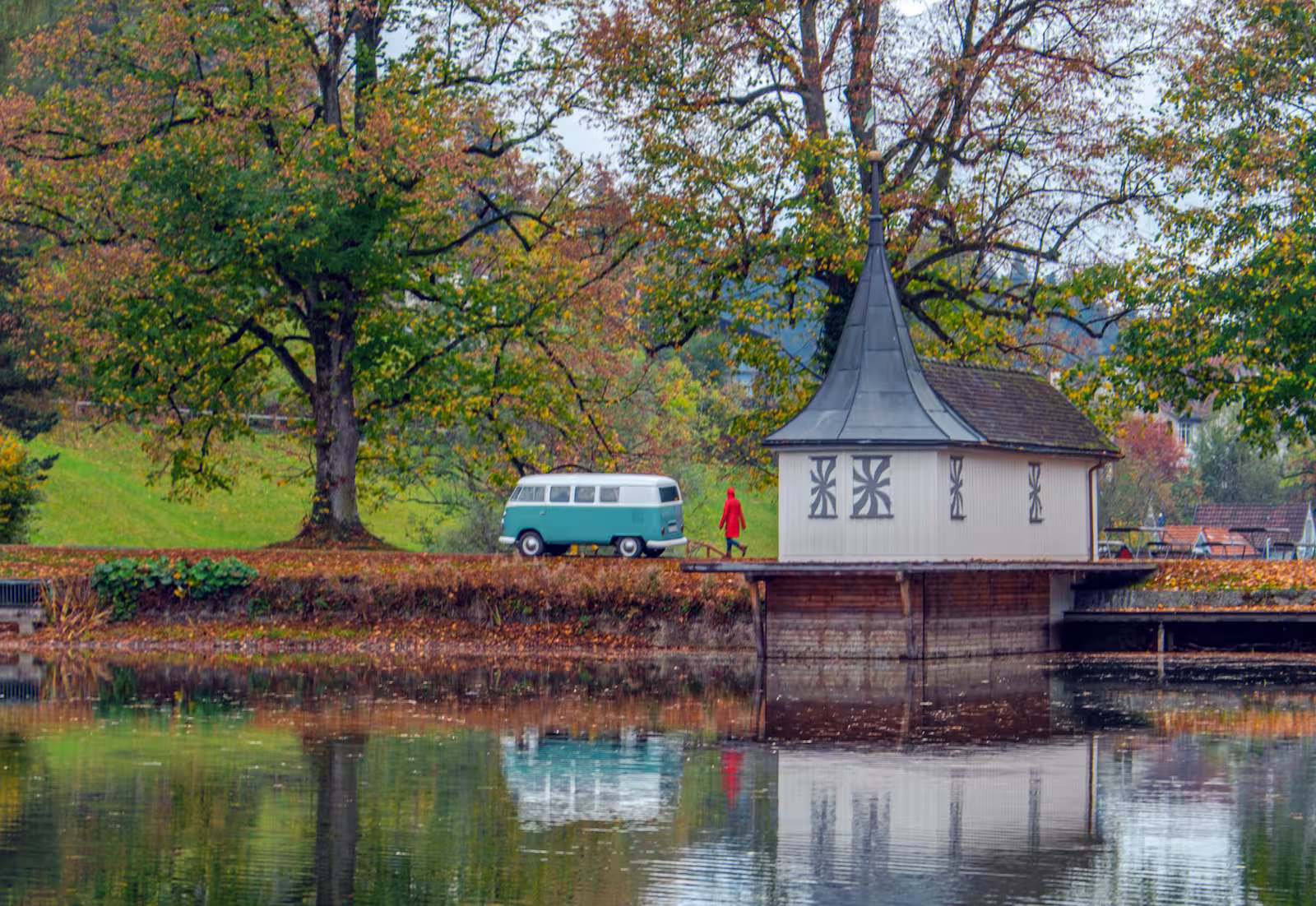Person in rotem Mantel läuft an einem blauen VW-Bus und einem kleinen Haus an einem herbstlichen Seeufer vorbei.